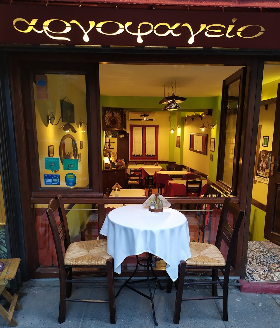 Η πρόσοψη του Αργοφαγείου τη μέρα — τραπεζάκι ντυμένο με λευκό τραπεζομάντιλο, δύο καρέκλες με ψάθα, η χρυσή επιγραφή «αργοφαγείο» στη βιτρίνα.
